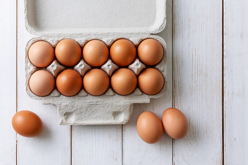 Open cardboard egg carton holding ten brown chicken eggs on white wooden surface with three eggs beside it