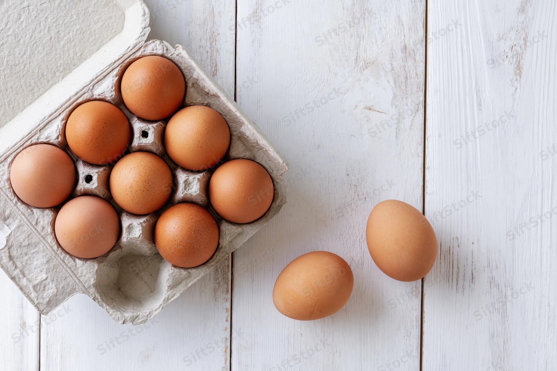 Open cardboard egg carton with brown chicken eggs on white wooden surface, two eggs beside it