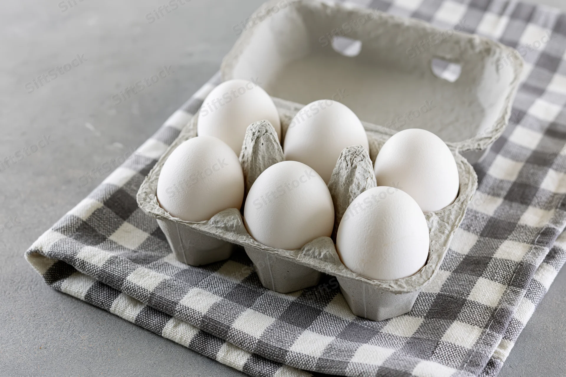 Open cardboard egg carton with six white eggs resting on a gray and white checkered cloth on a flat surface