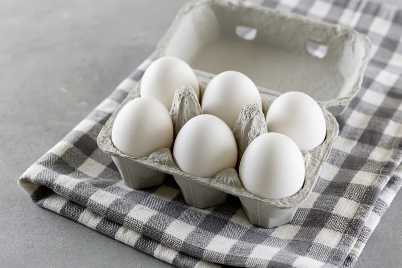 Open cardboard egg carton with six white eggs resting on a gray and white checkered cloth on a flat surface