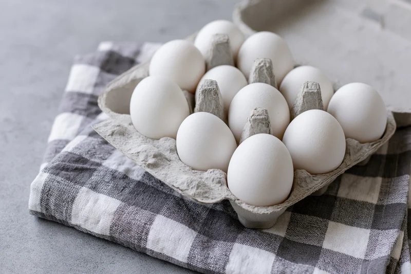 Open cardboard egg carton filled with white eggs resting on a checkered cloth on a kitchen surface