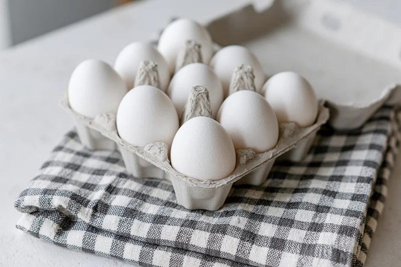 Open cardboard egg carton filled with white eggs sitting on a checkered cloth on a kitchen countertop