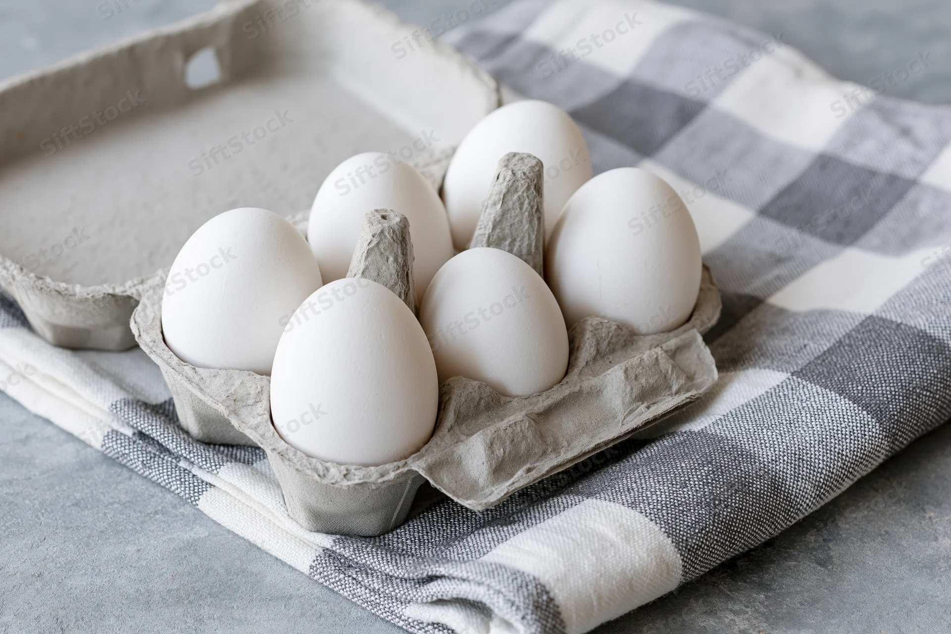 White eggs in open cardboard carton resting on checkered cloth on countertop