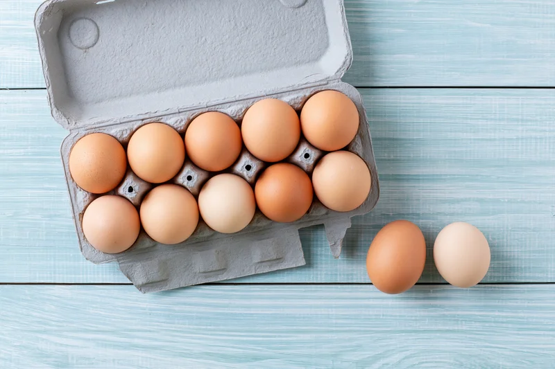 Open cardboard egg carton holding ten brown chicken eggs on a blue wooden table with two eggs beside it