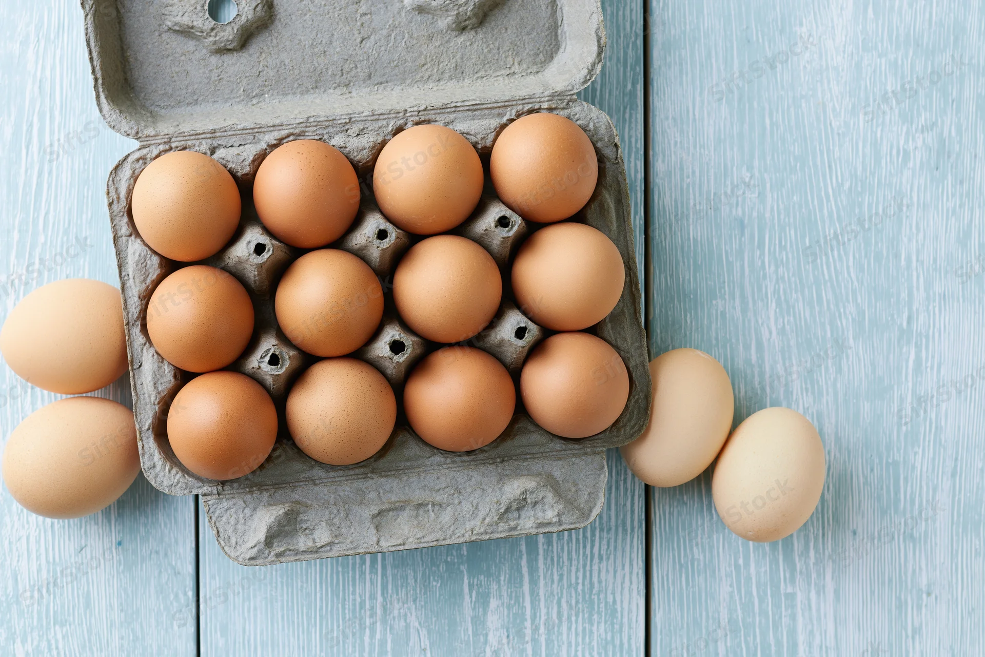 Open cardboard egg carton holding ten brown chicken eggs on a blue wooden surface with additional eggs scattered around