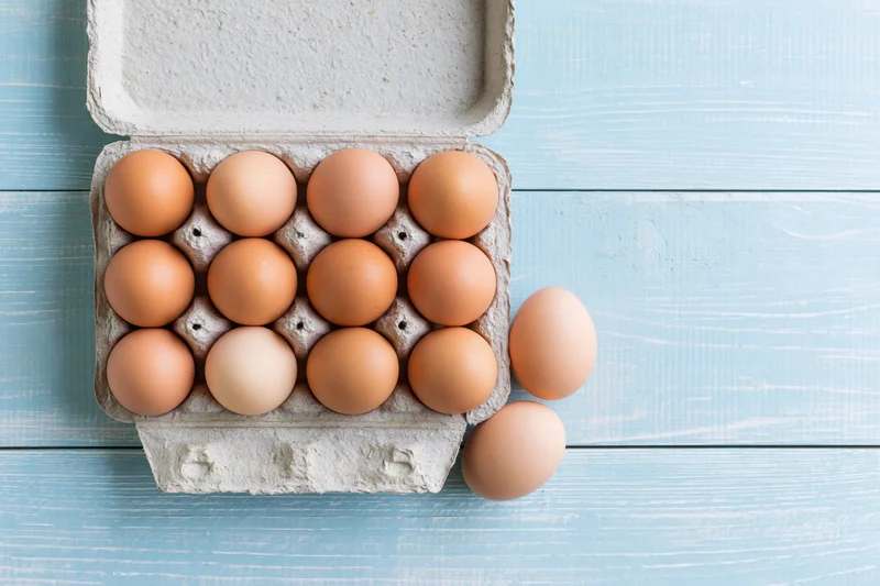 Open cardboard egg carton holding ten brown chicken eggs on blue wooden surface with two eggs beside it