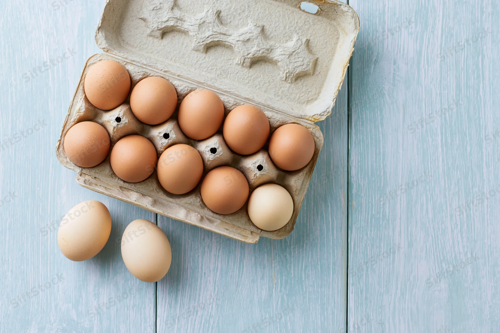 Open cardboard egg carton holding ten brown chicken eggs on a blue wooden surface with two eggs beside it