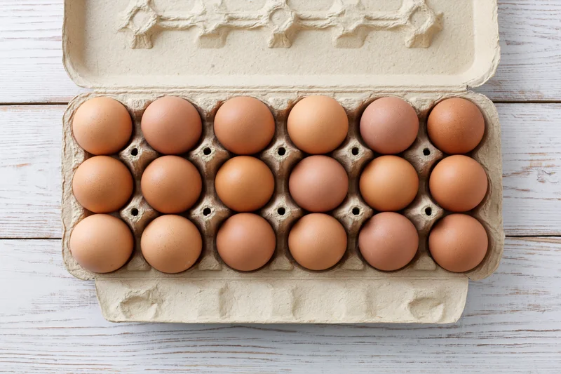 Open cardboard egg carton holding twelve brown eggs on white wooden surface