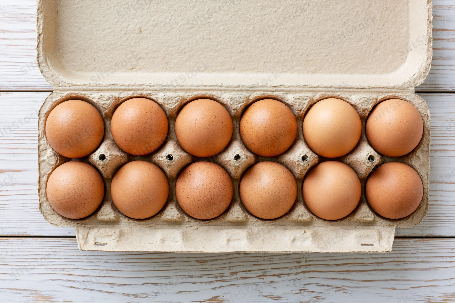 Twelve brown eggs arranged in an open cardboard carton on a white wooden surface