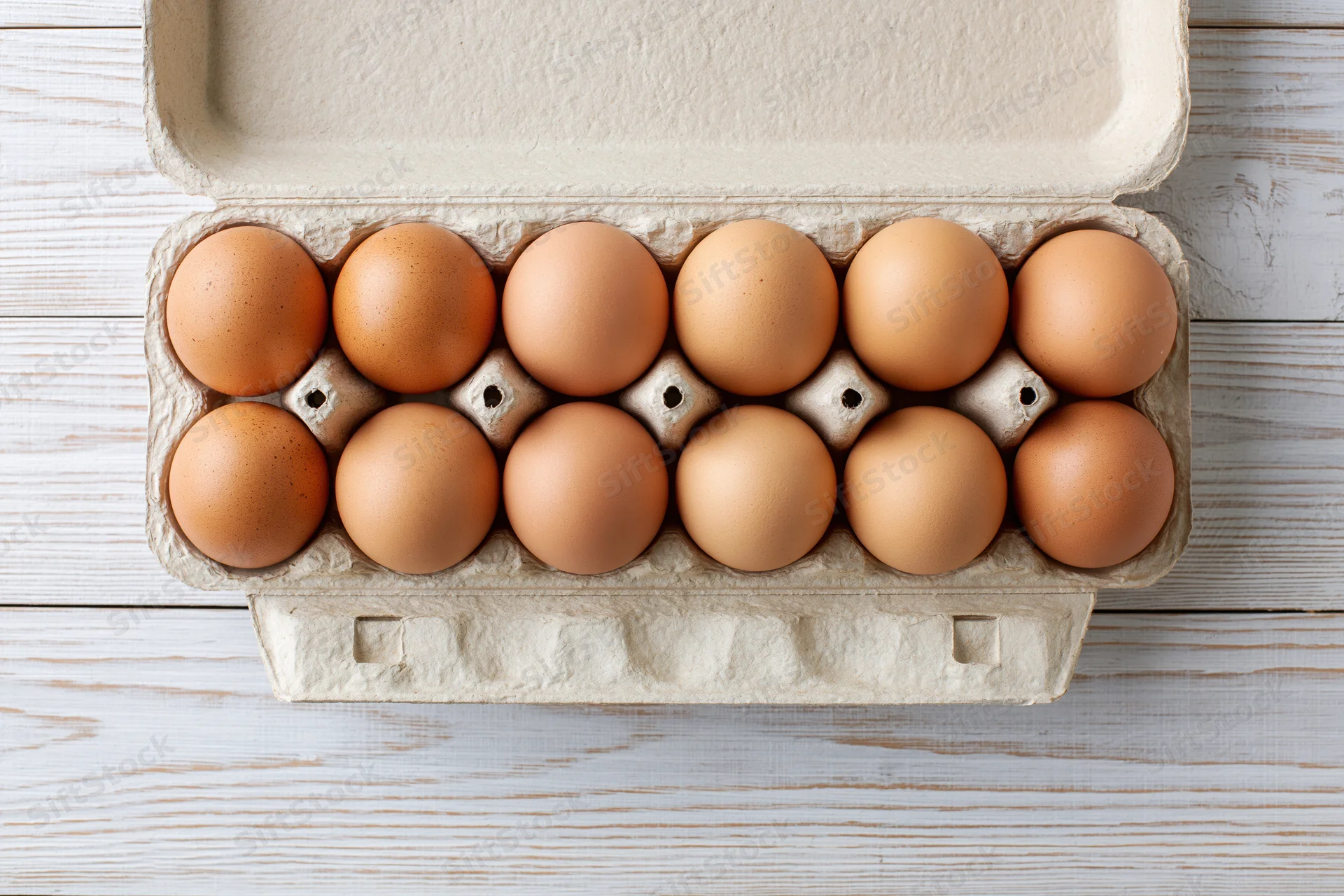 Open cardboard egg carton holding twelve brown eggs on a white wooden surface