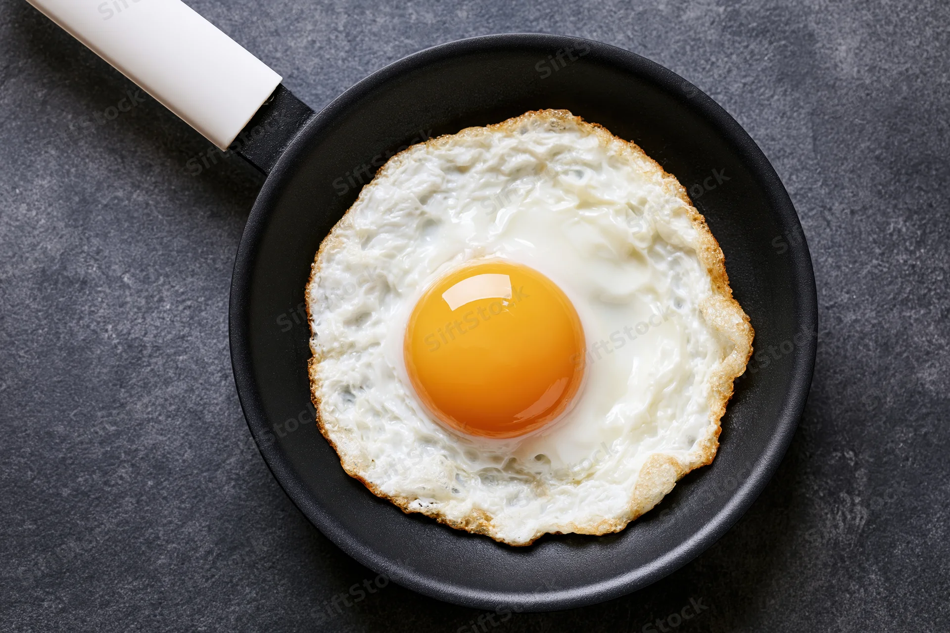 Single fried egg in a small black nonstick pan viewed from above on dark surface