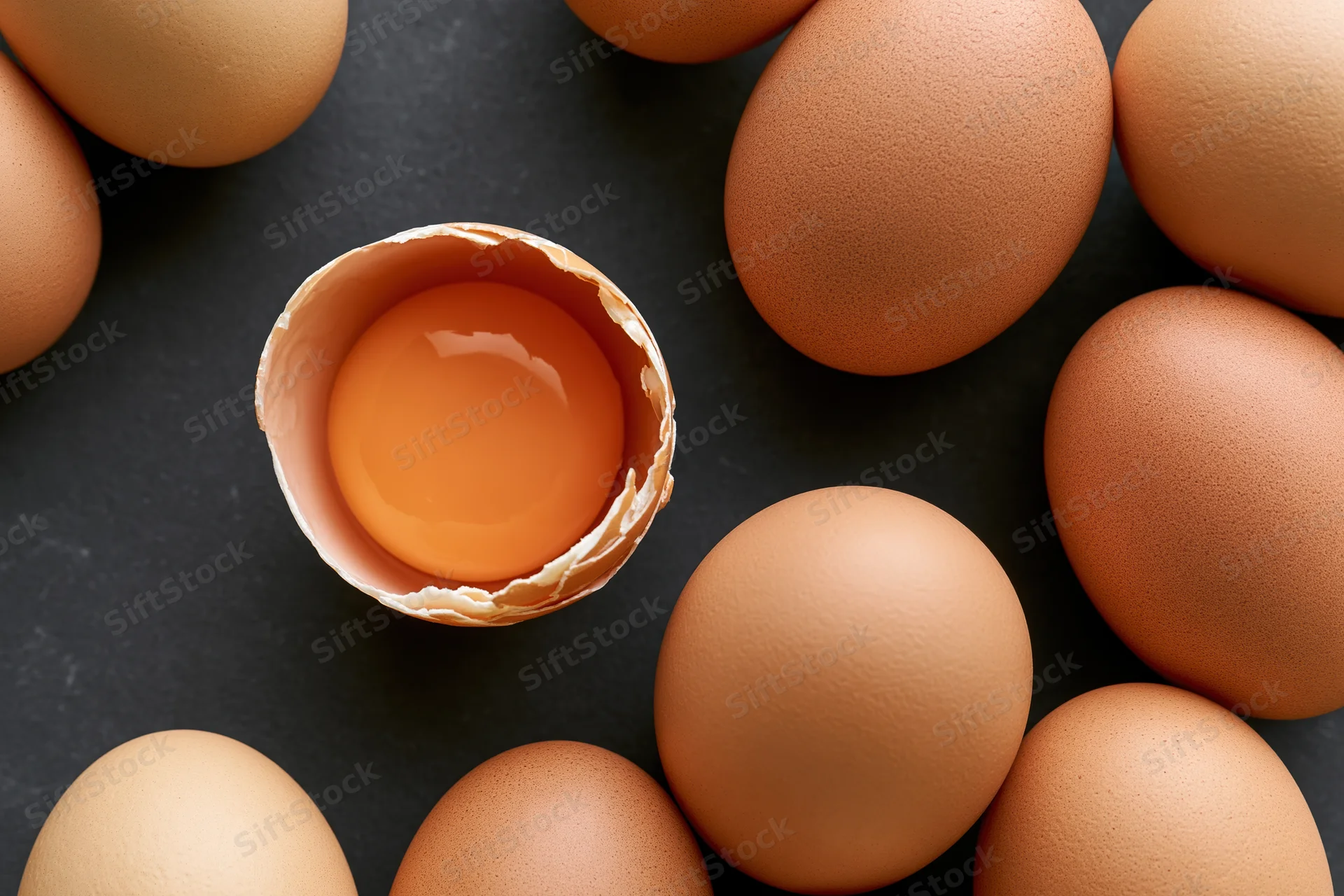 Brown chicken eggs arranged in flat lay with one cracked open showing yolk on dark surface
