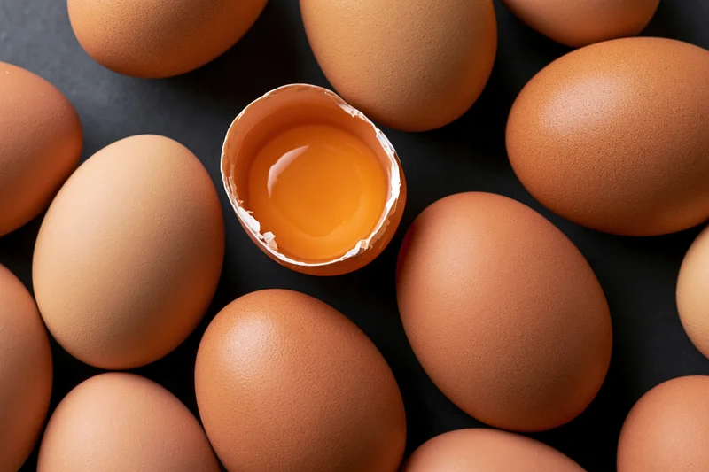 Brown chicken eggs arranged in flat lay with one cracked open showing yolk on dark surface