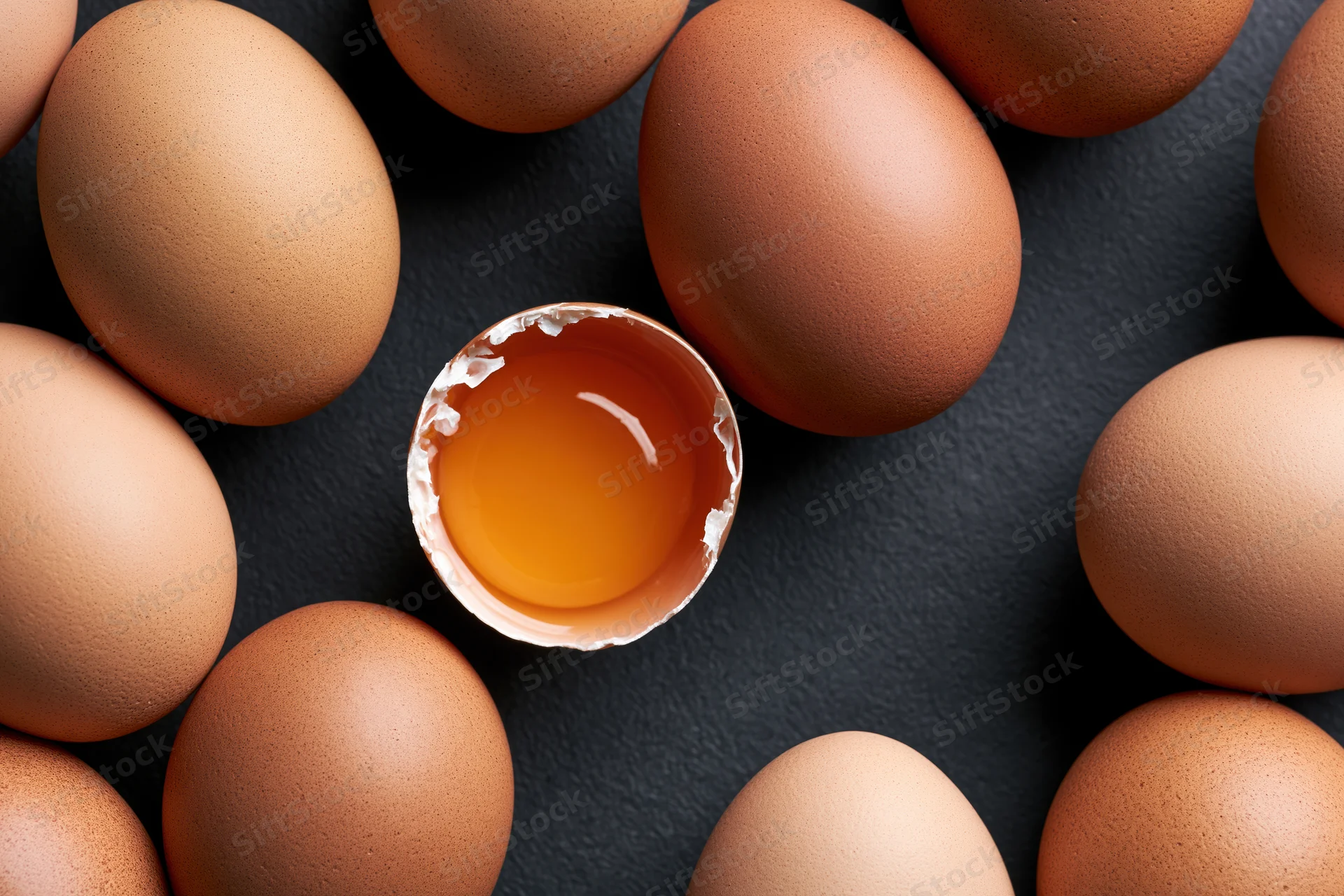 Brown chicken eggs arranged in circle with one cracked open showing yolk on dark surface