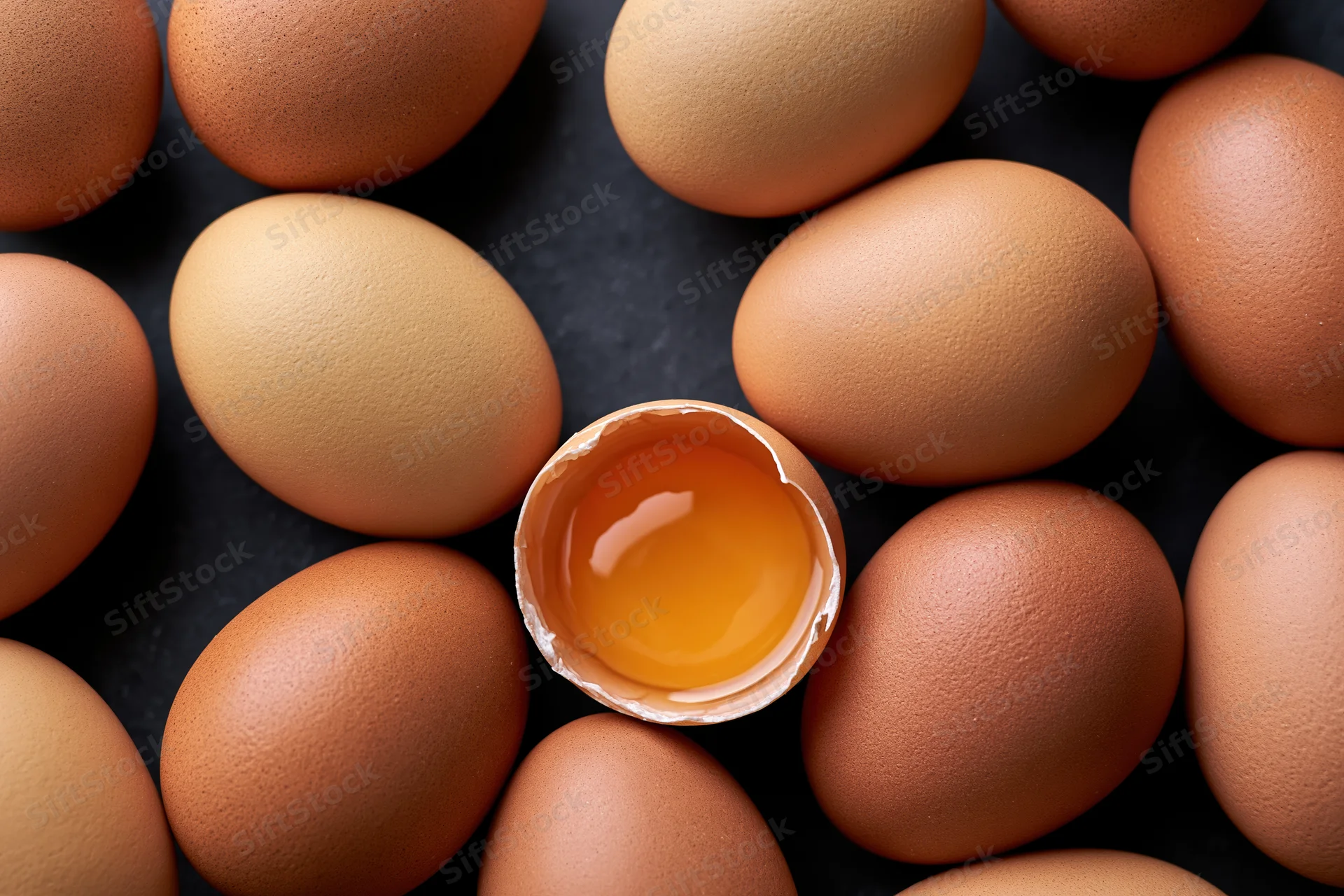 Overhead flat-lay of brown chicken eggs with one cracked open revealing yolk on dark surface