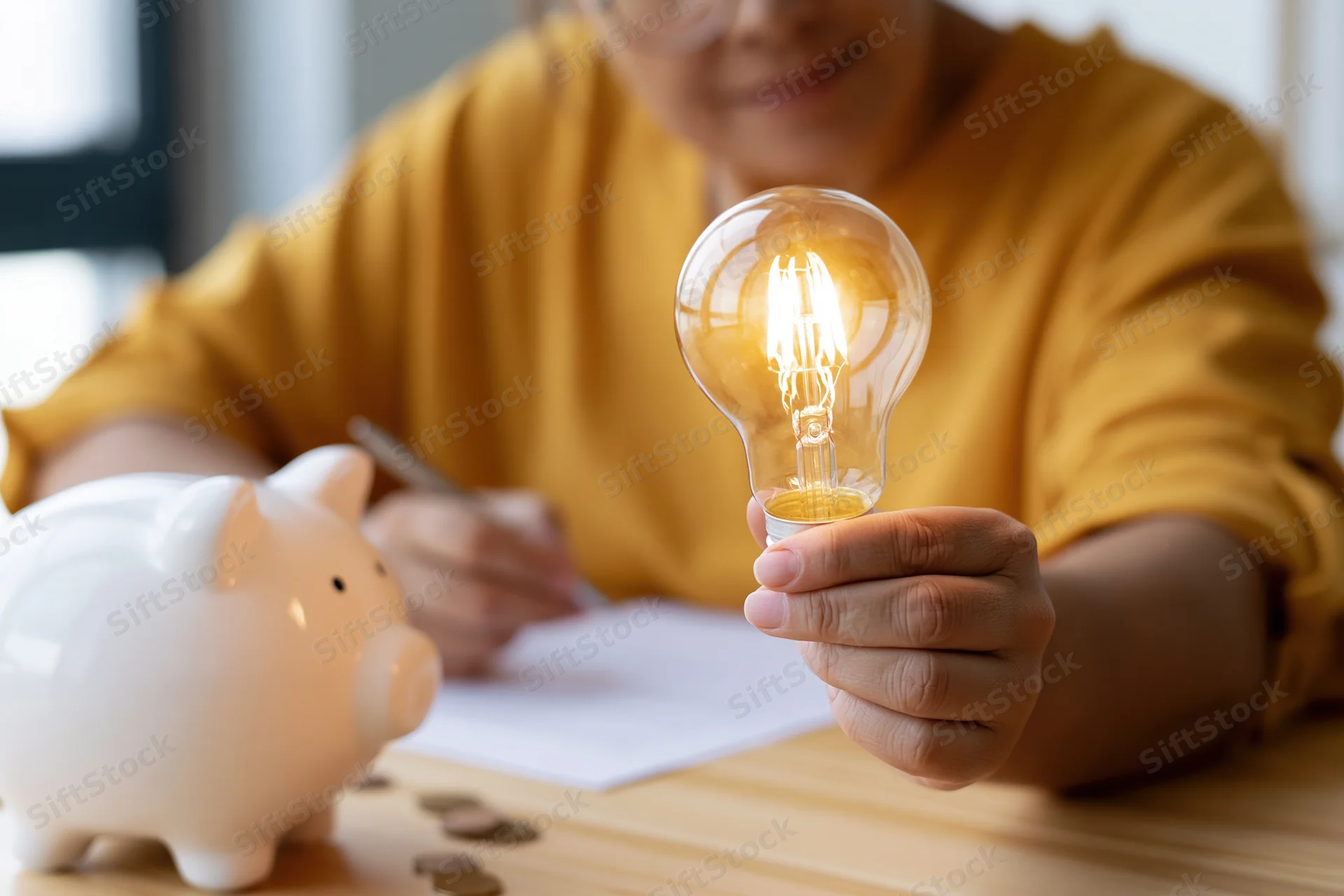 Woman holding glowing incandescent lightbulb while writing on paper at wooden desk with piggybank and coins nearby