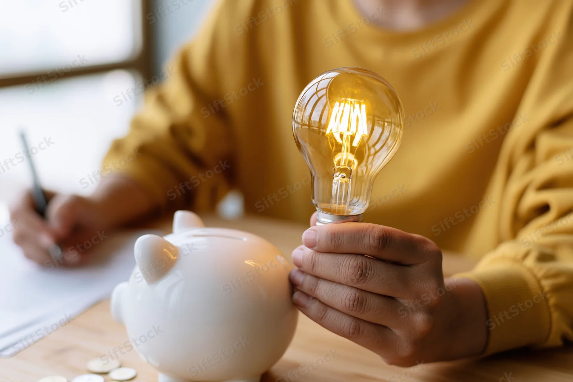 Person in yellow shirt holding glowing incandescent lightbulb while writing on paper at wooden desk with piggybank and coins nearby