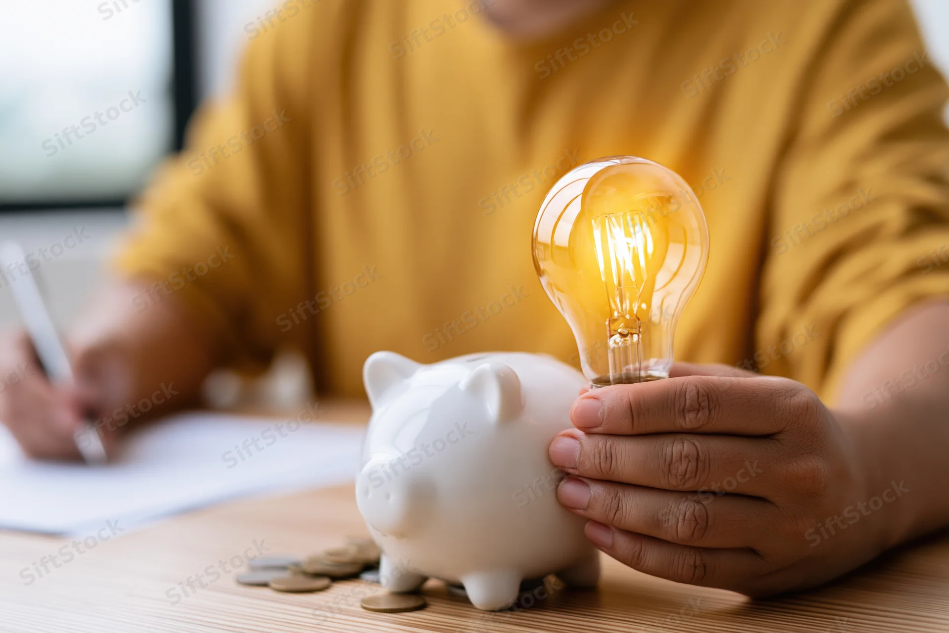Person holding glowing lightbulb over piggybank while writing on paper at desk