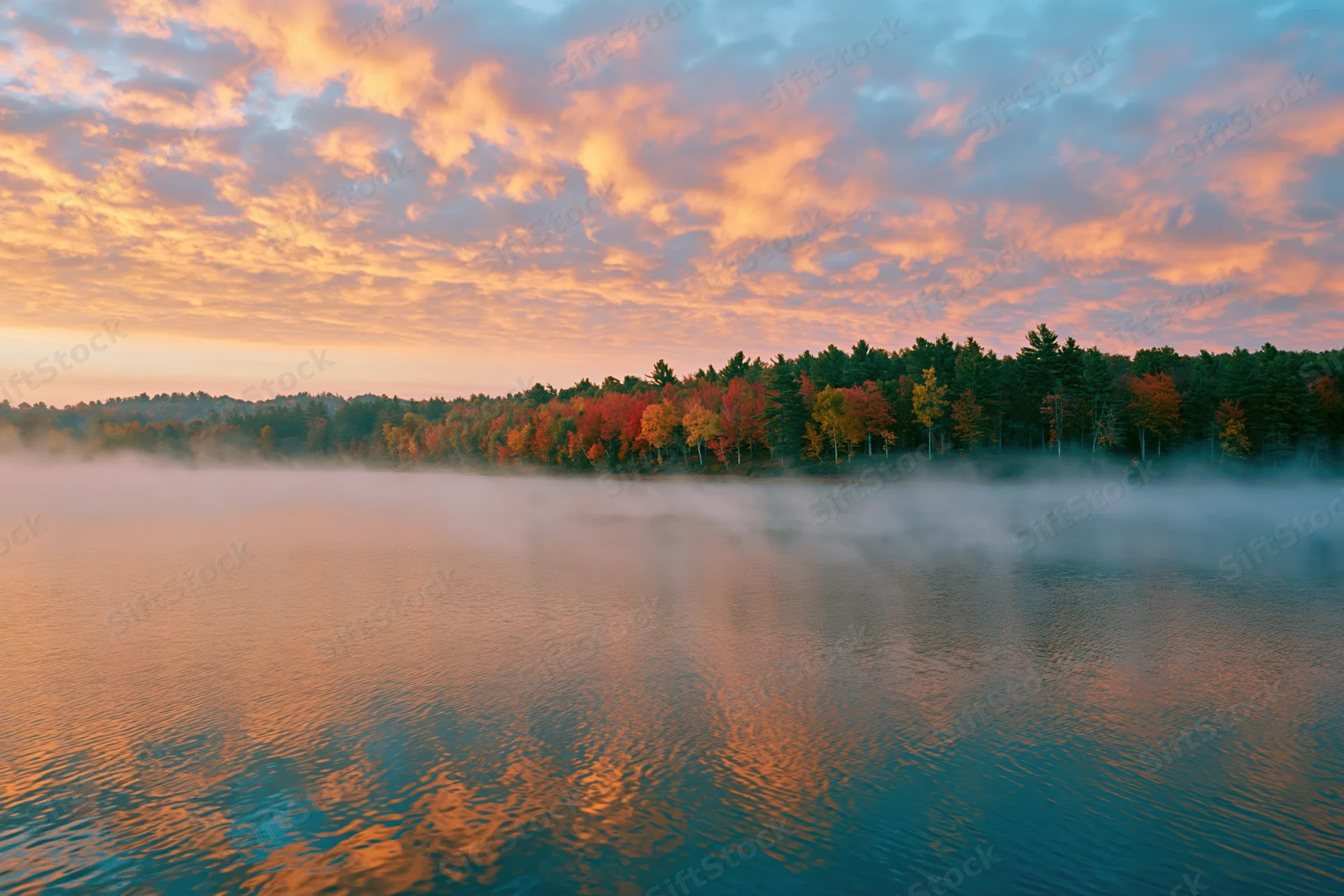 Calm lake reflecting autumn trees at sunrise with mist drifting over water
