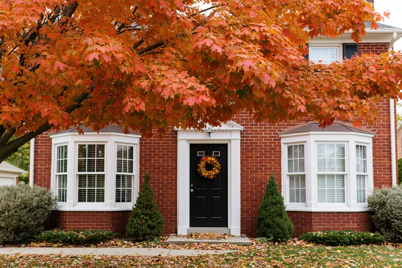 Red brick colonial house facade with black front door and autumn wreath under maple tree with orange leaves