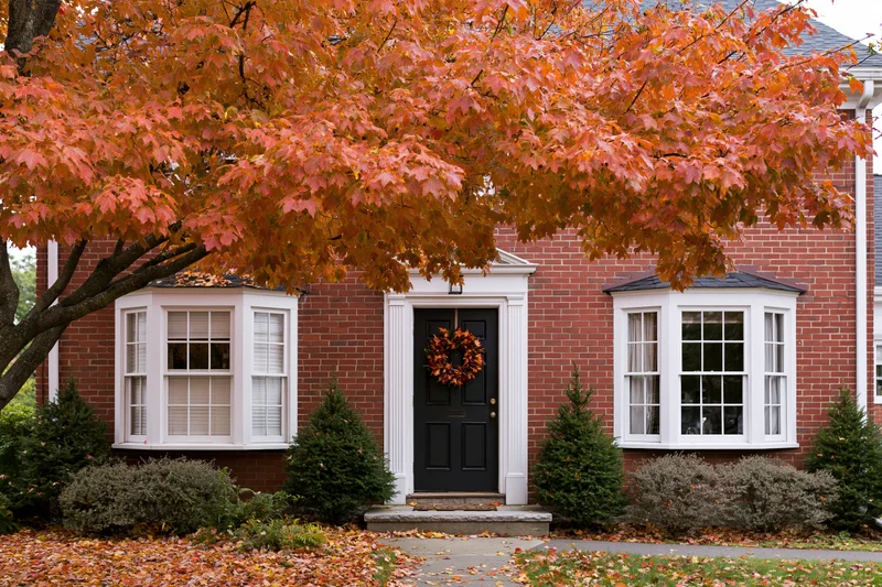 Red brick colonial house facade with black front door decorated with autumn wreath and surrounded by fall foliage