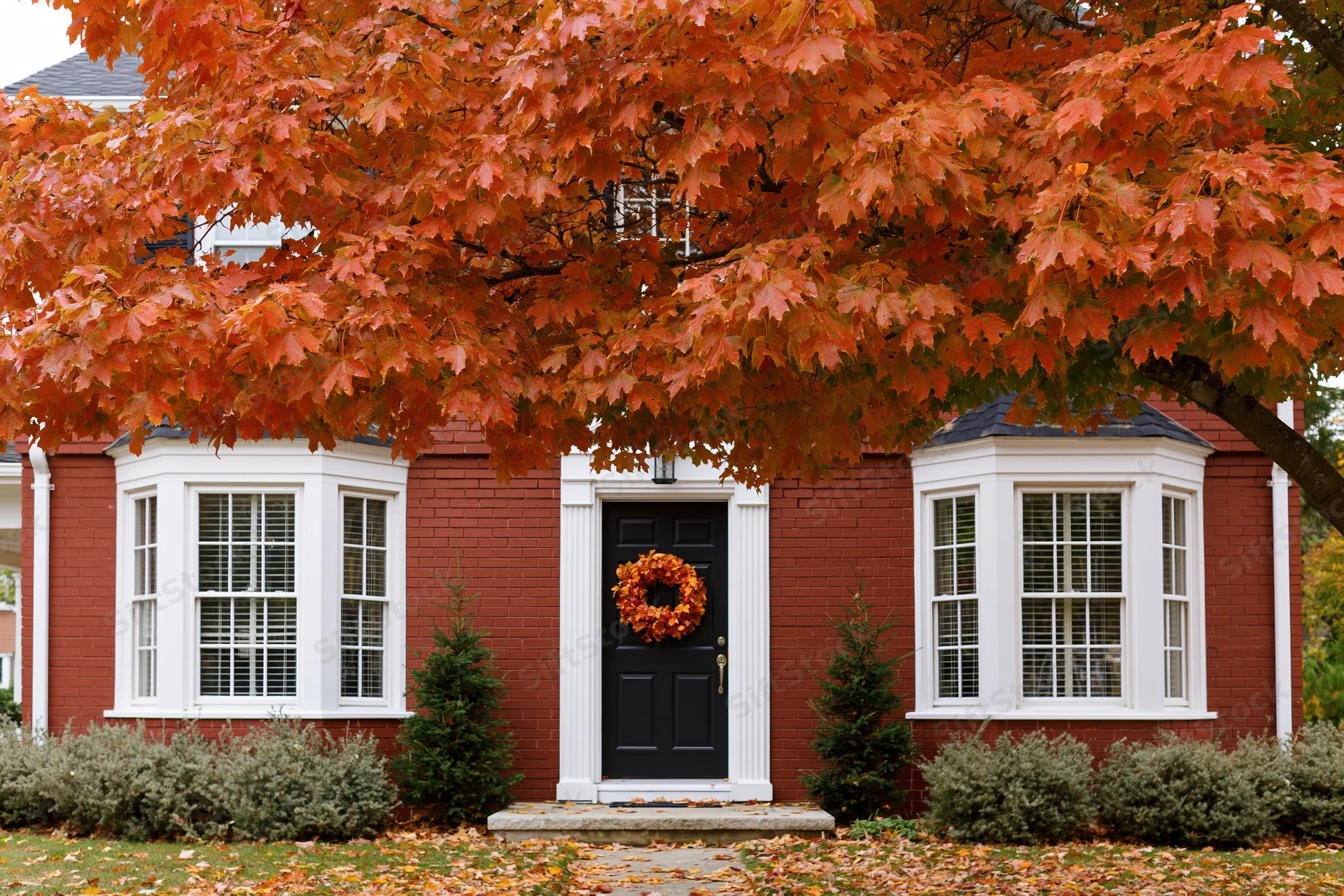 Red brick colonial house facade with black front door and autumn wreath under maple tree with orange leaves