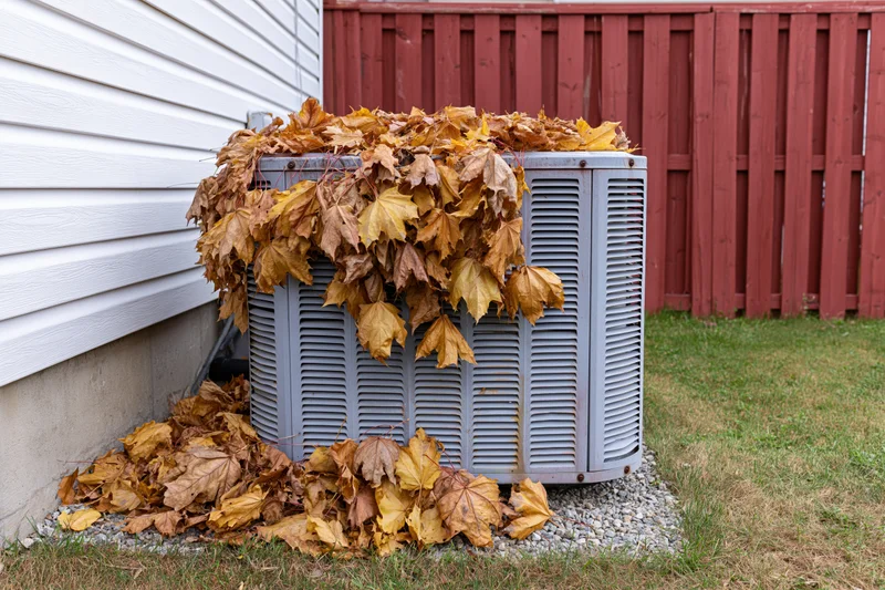 Outdoor air conditioning condenser unit covered in fallen autumn leaves in a residential backyard