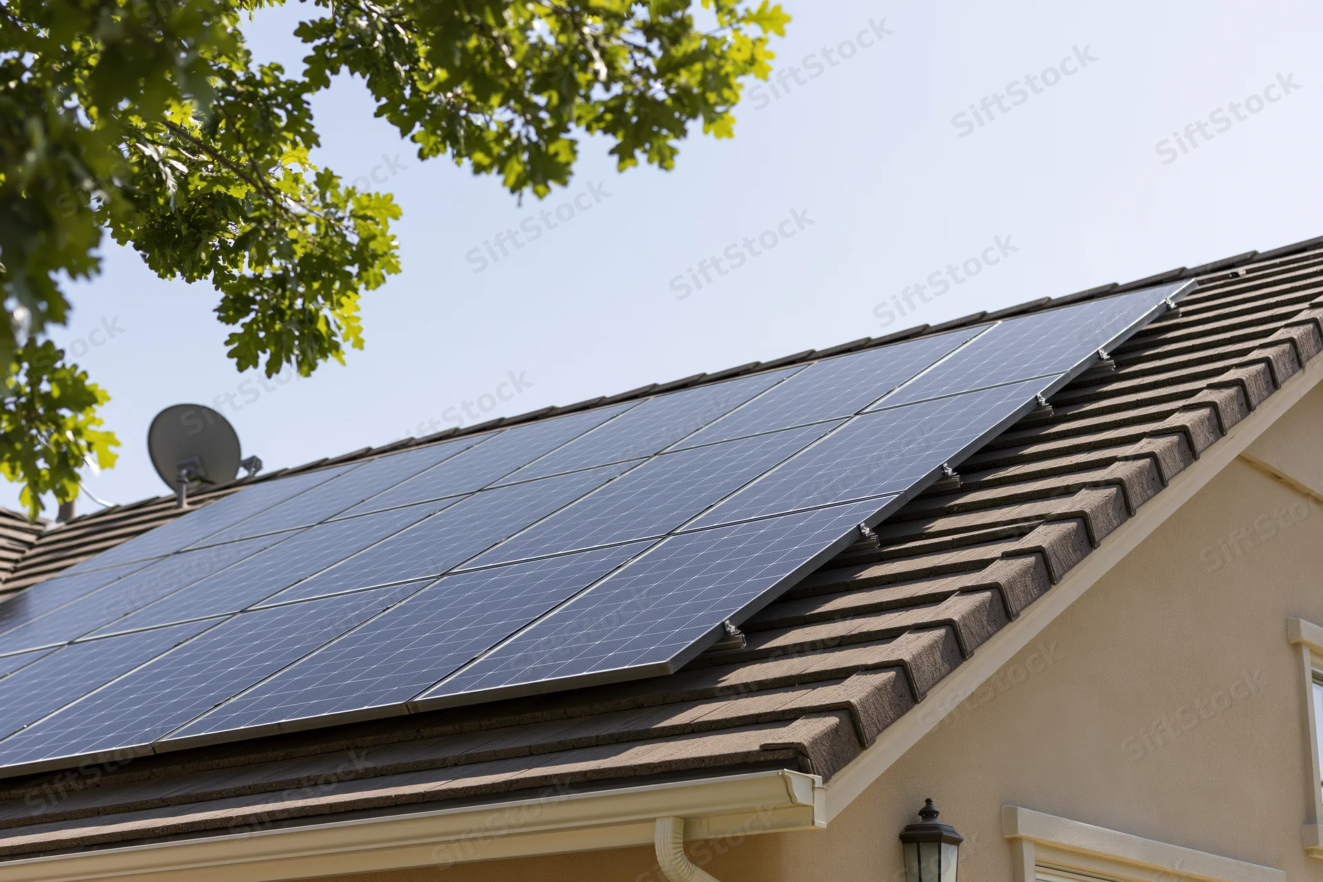 Solar panels installed in a grid on a residential rooftop with shingles and a satellite dish under a clear sky