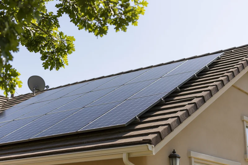 Solar panels installed in a grid on a residential rooftop with shingles and a satellite dish under a clear sky