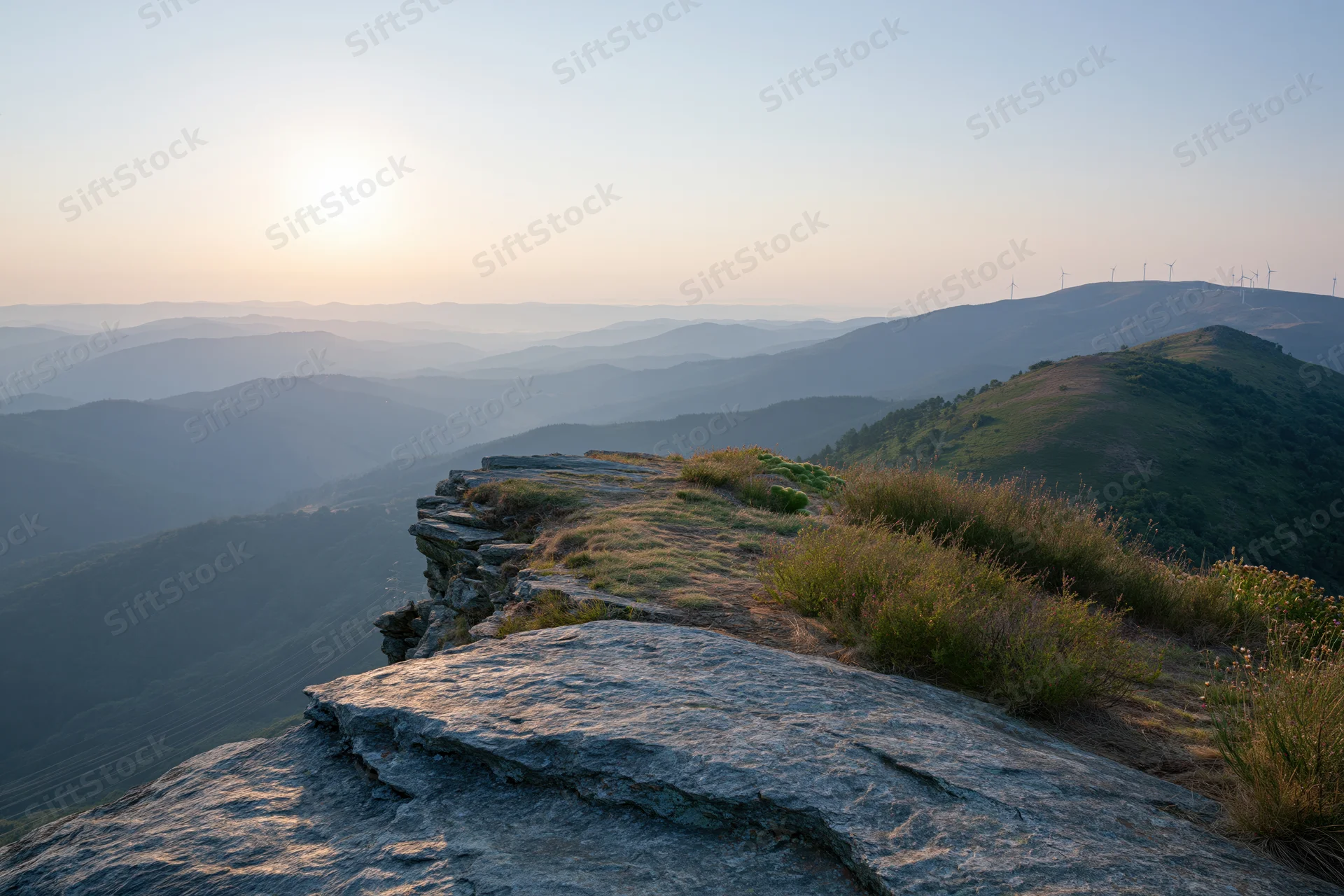 Rocky cliff edge with sparse grass and shrubs overlooking layered mountains at sunset