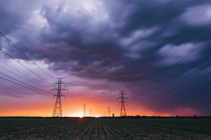 Row of high-voltage electricity pylons stretching across a flat rural field at sunset under stormy clouds