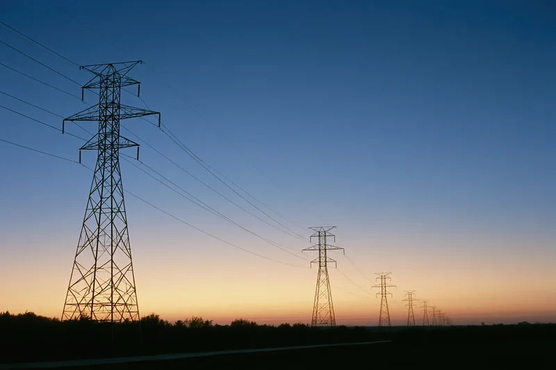 Row of high-voltage electricity transmission towers receding into the distance at dusk over a rural landscape
