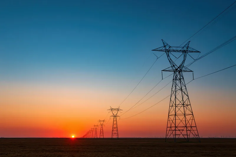 Row of high-voltage electricity transmission towers silhouetted against a sunset sky over a flat rural field