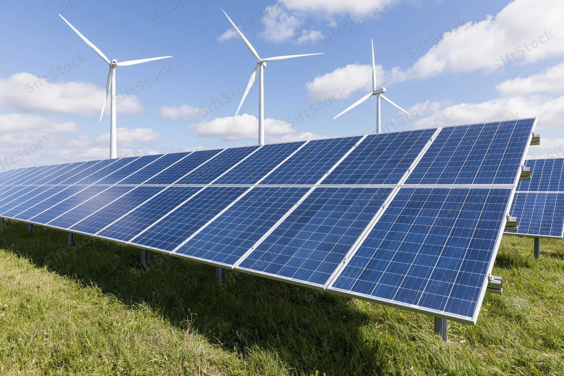 Rows of blue photovoltaic solar panels installed in a green field with wind turbines in the background under a partly cloudy sky