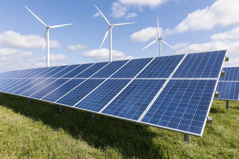Rows of blue photovoltaic solar panels installed in a green field with wind turbines in the background under a partly cloudy sky