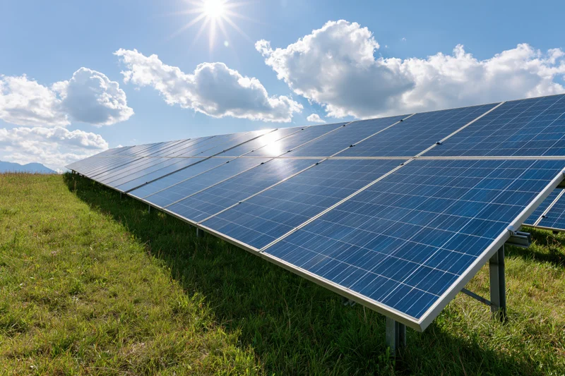 Rows of blue photovoltaic solar panels installed on a grassy field under a bright sunlit sky with scattered clouds