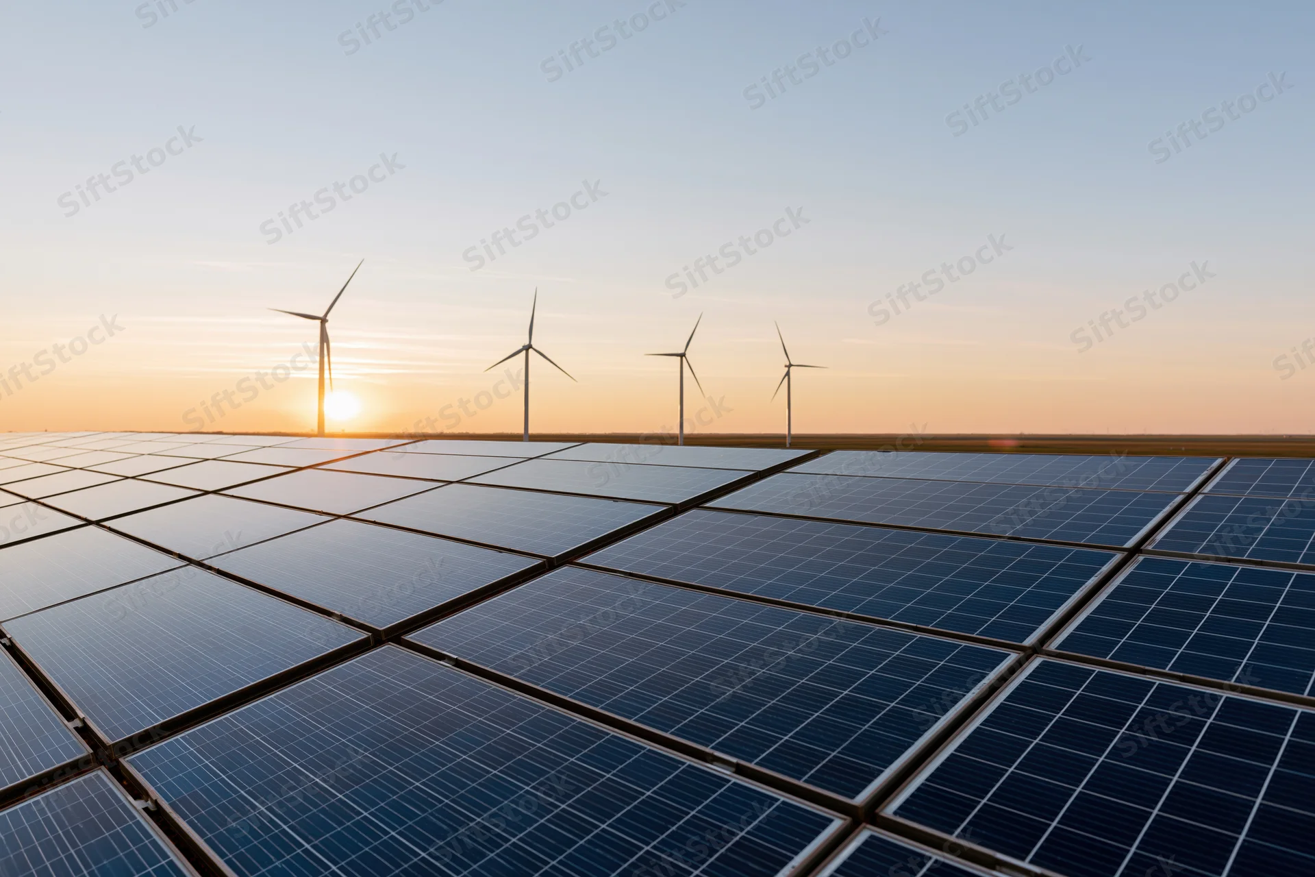 Rows of blue solar panels in a field with wind turbines at sunset