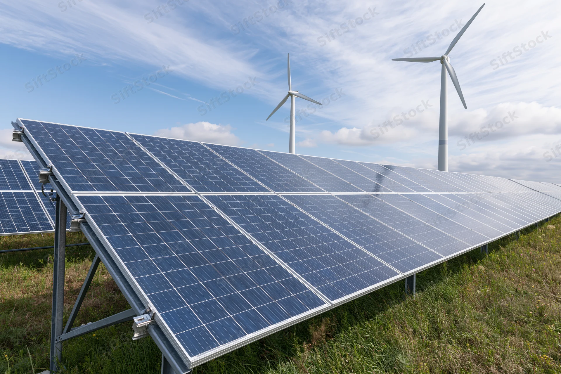 Rows of blue solar panels mounted on metal frames in an open grassy field with wind turbines in the background under a partly cloudy sky