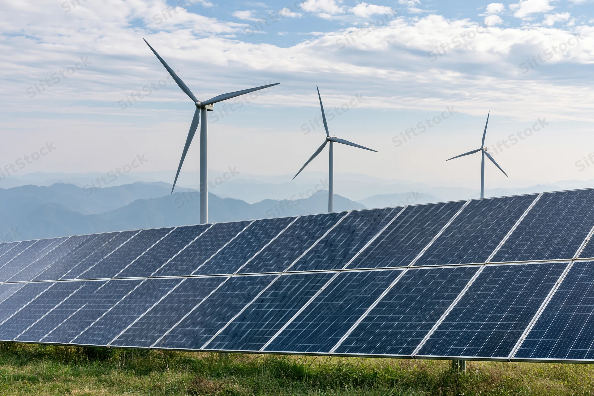Rows of dark blue photovoltaic solar panels in a green field with wind turbines and mountains in the background