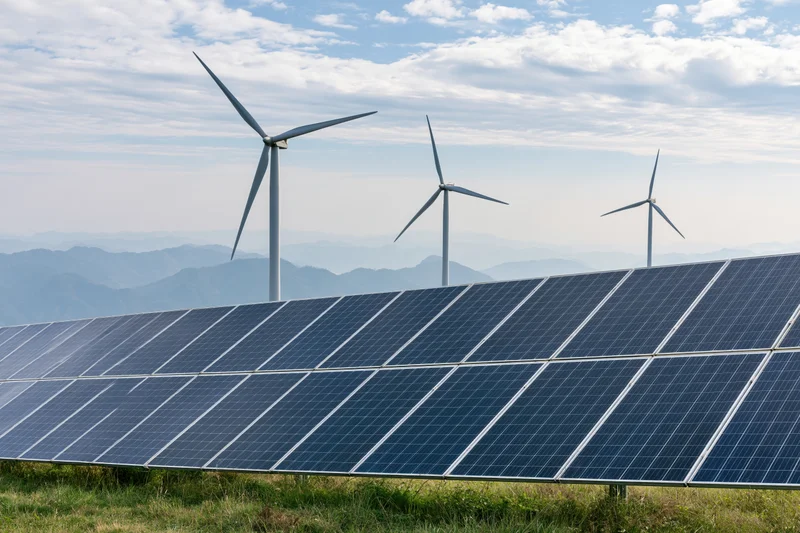 Rows of dark blue photovoltaic solar panels in a green field with wind turbines and mountains in the background
