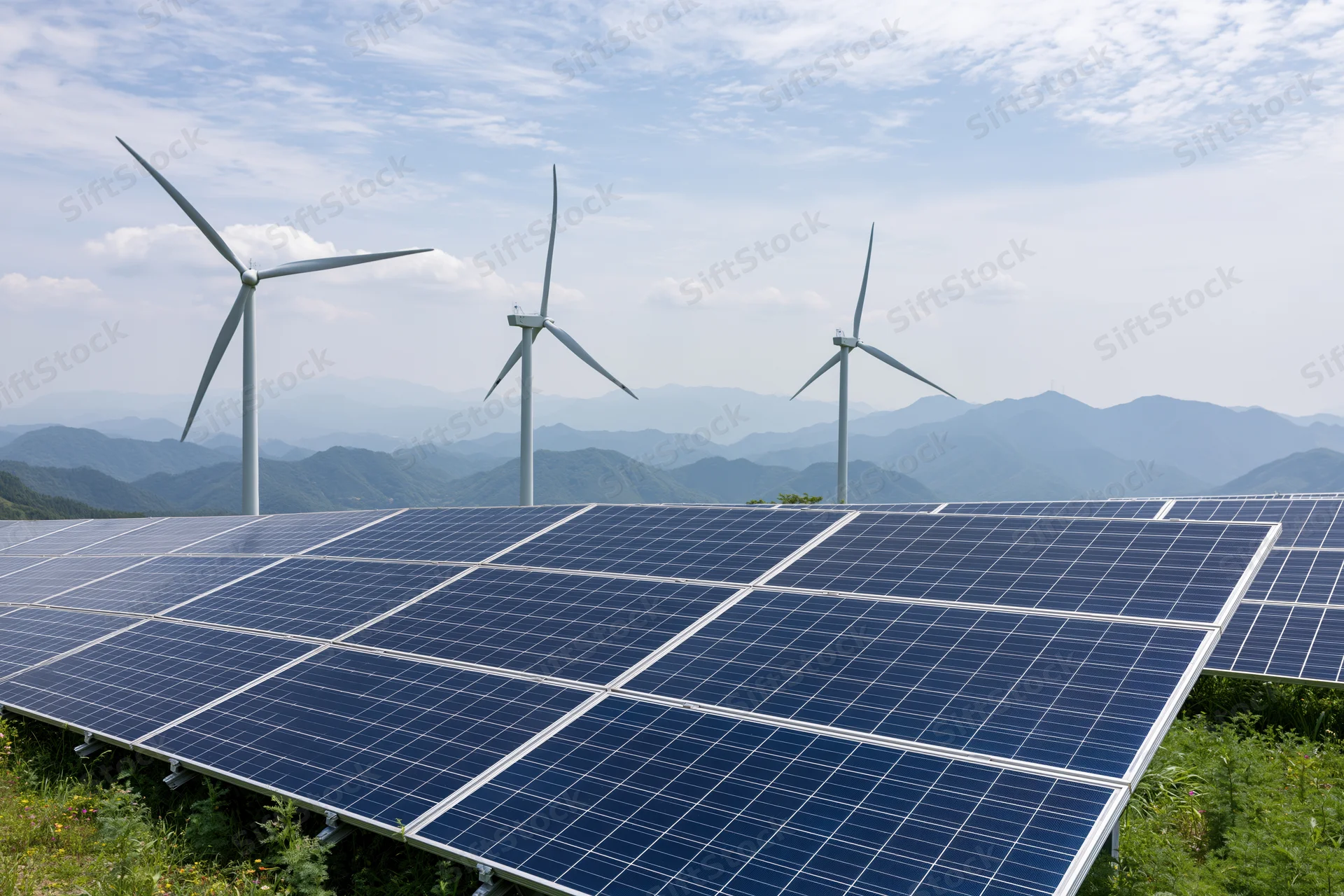 Rows of dark blue photovoltaic solar panels in a green field with wind turbines and mountains in the background