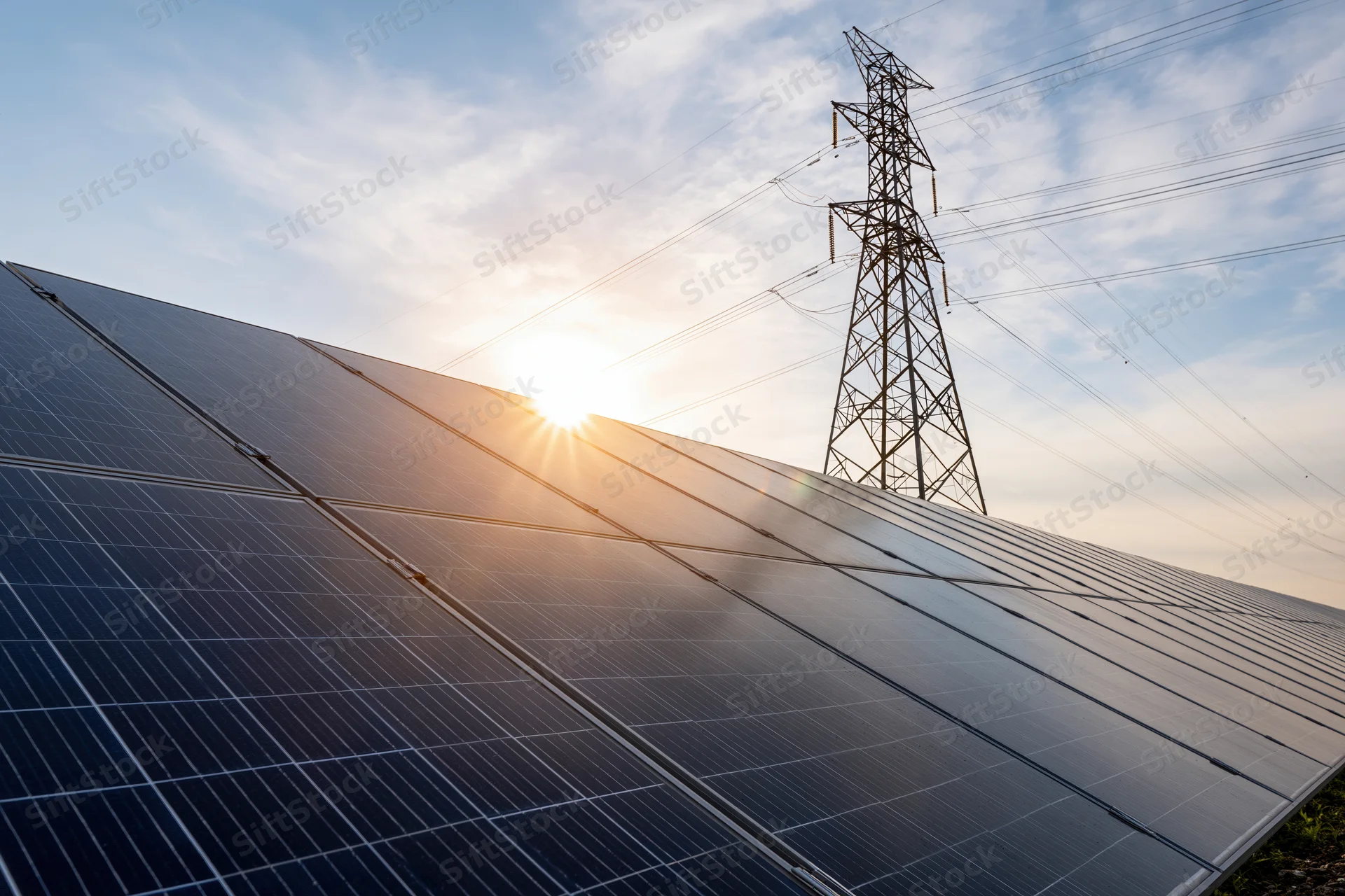 Rows of dark blue solar panels stretching across a field with a transmission tower and sunlit sky in the background