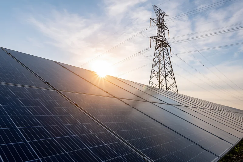 Rows of dark blue solar panels stretching across a field with a transmission tower and sunlit sky in the background