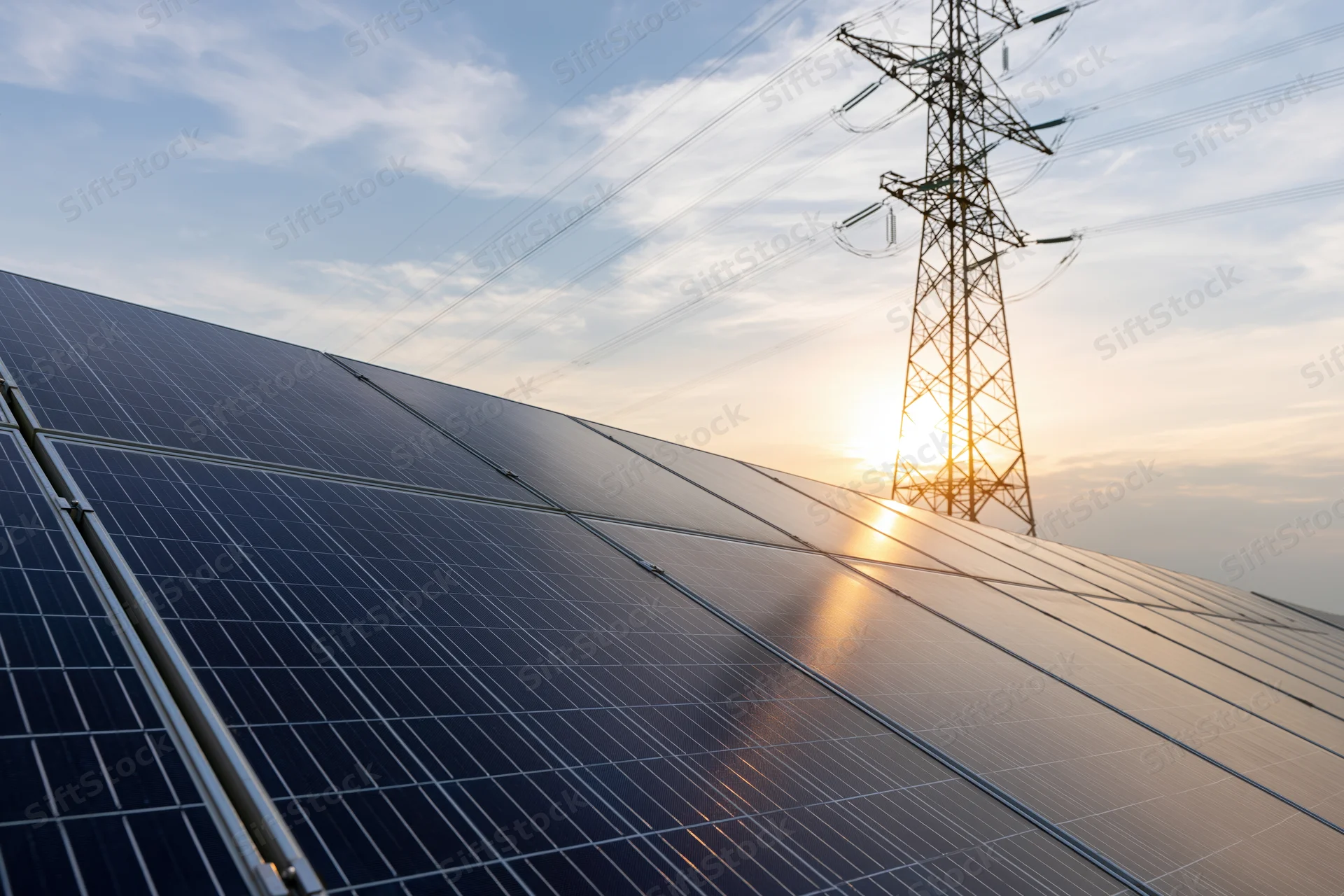 Rows of dark blue solar panels aligned on a sloped surface with a transmission tower and power lines in the background during sunset