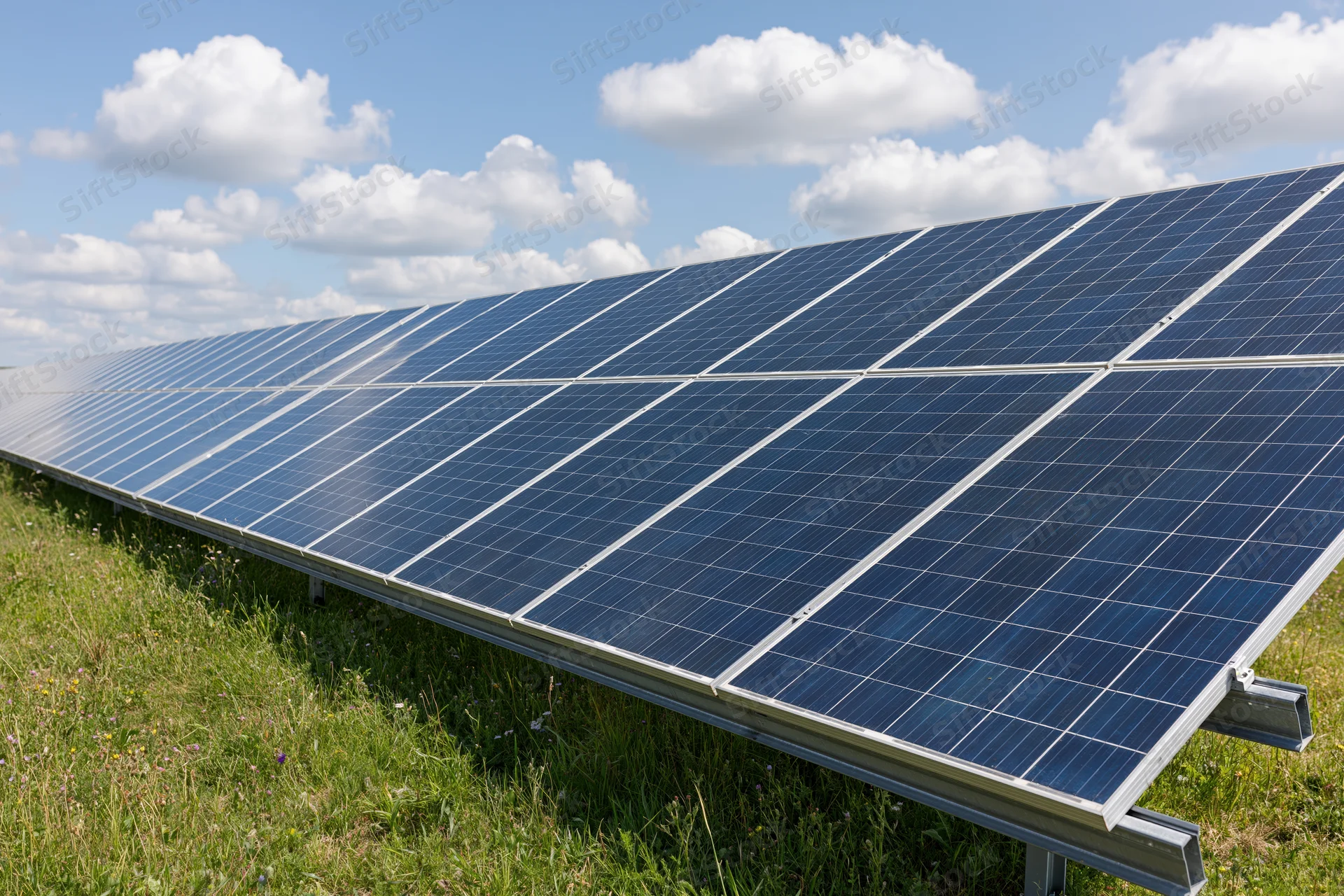 Rows of ground-mounted solar panels stretching across a green field under a blue sky with white clouds