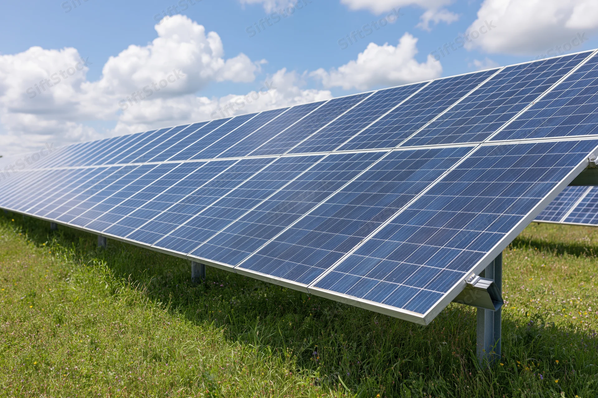 Rows of ground-mounted solar panels stretching across a green field under a blue sky with white clouds