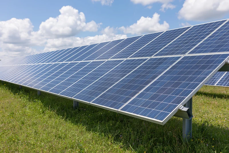 Rows of ground-mounted solar panels stretching across a green field under a blue sky with white clouds