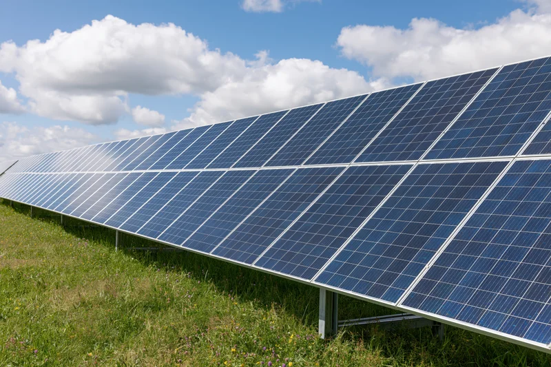 Rows of ground-mounted solar panels stretching across a green field under a blue sky with white clouds