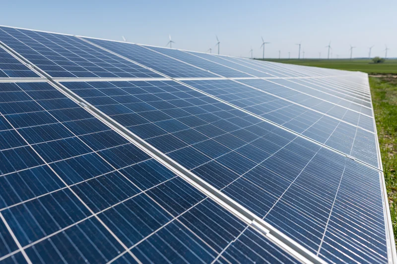 Rows of solar panels angled toward the sky in a utility-scale field with wind turbines in the distance
