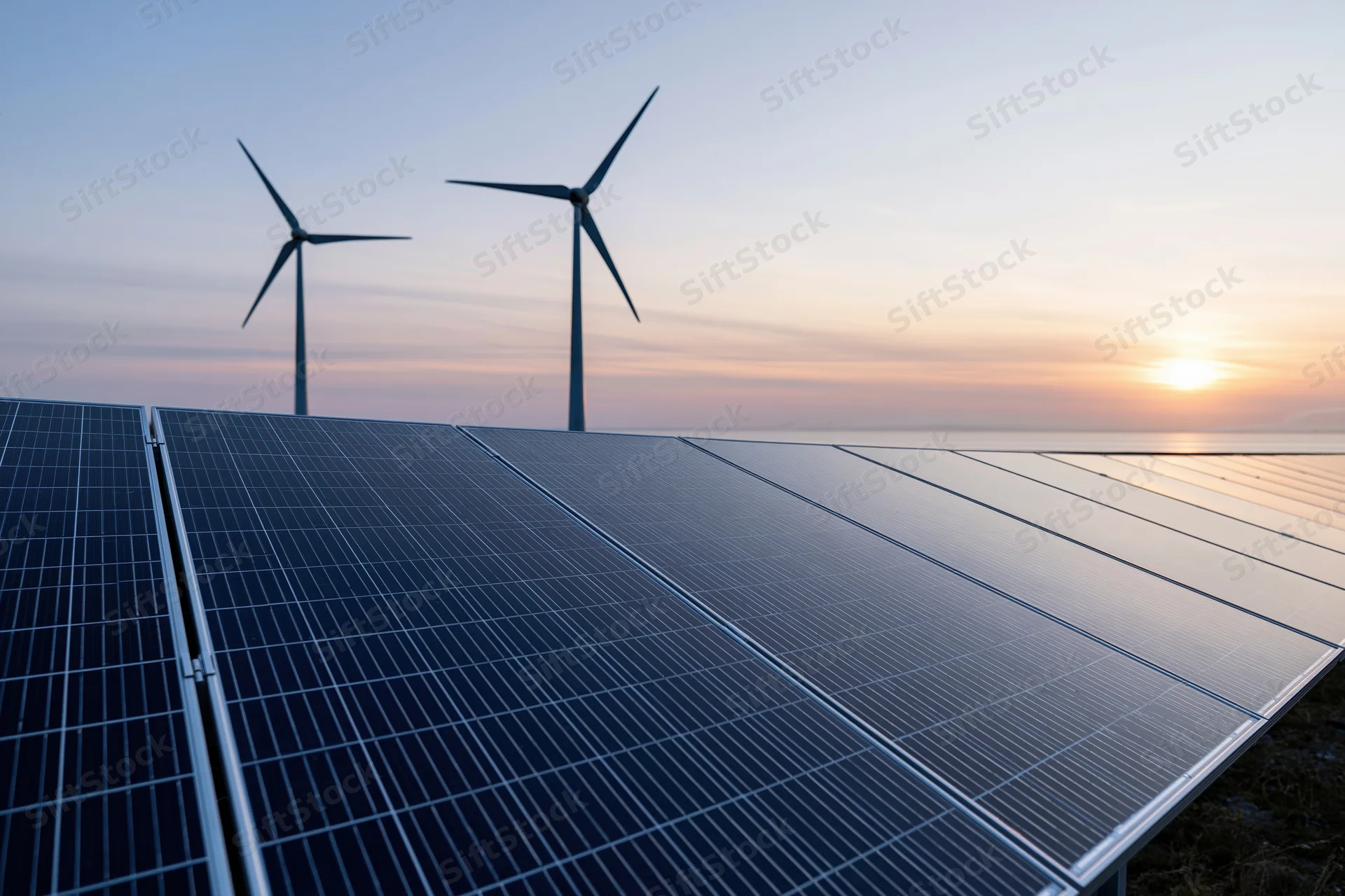 Rows of solar panels in foreground with wind turbines in background at sunset near ocean